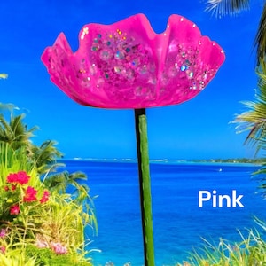 Pu&ograve; includere: Un fiore di vetro rosa con dettagli glitterati, su uno sfondo di spiaggia tropicale. Il fiore &egrave; su uno stelo di metallo verde. Il testo "Pink" &egrave; visibile nell'angolo in basso a destra dell'immagine.
