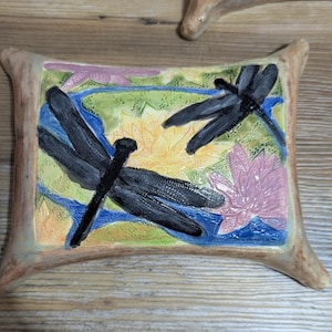 May include: A ceramic tile with a raised design of two dragonflies and water lilies in shades of blue, yellow, pink, and green.