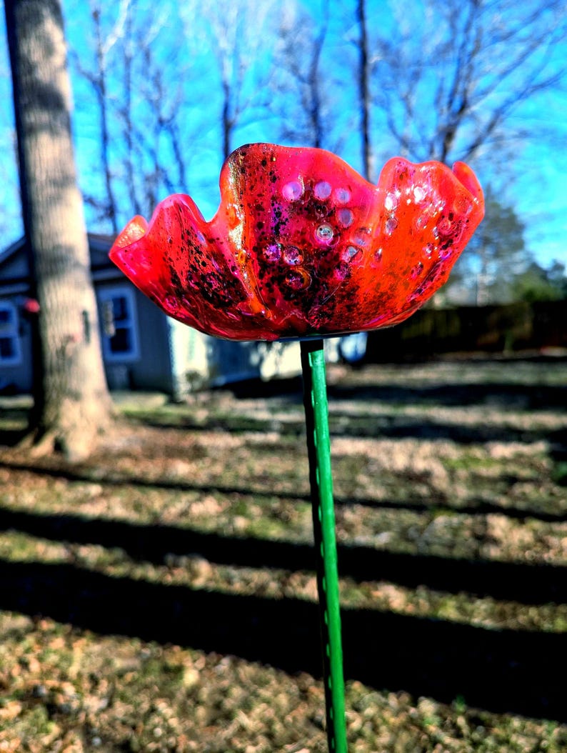 Pu&ograve; includere: Un fiore di papavero di vetro rosso con uno stelo verde. Il fiore ha una superficie testurizzata e una finitura scintillante.