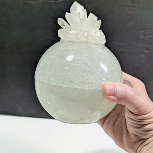 May include: A round, iridescent, glitter-filled orb with a cluster of clear crystals on top. The orb is held in a hand, showcasing its spherical shape and sparkling interior. The crystals add a touch of elegance.
