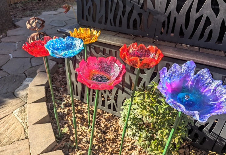 Pu&ograve; includere: Cinque sculture di fiori in vetro colorato con steli in metallo verde. I fiori sono in tonalit&agrave; di blu, rosso, rosa e arancio. I fiori sono disposti in un giardino.