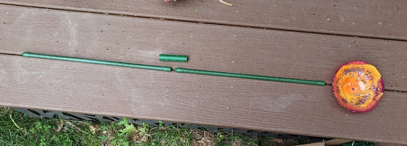 Pu&ograve; includere: Un'asta di metallo verde &egrave; rotta in tre pezzi. L'asta &egrave; appoggiata su una superficie di legno marrone. Un oggetto decorativo rotondo, arancione e giallo si trova sul lato destro dell'immagine.