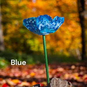 Pu&ograve; includere: Un fiore di vetro blu con vortici bianchi, su uno stelo verde, in piedi su un mucchio di foglie autunnali. Il fiore &egrave; un pezzo unico e decorativo per un giardino o una casa.