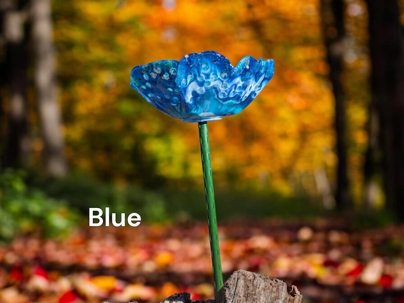 Pu&ograve; includere: Un fiore di vetro blu con vortici bianchi, su uno stelo verde, in piedi su un mucchio di foglie autunnali. Il fiore &egrave; un pezzo unico e decorativo per un giardino o una casa.