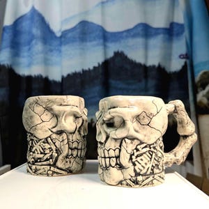 May include: Two ceramic mugs with a skull design. The mugs are off-white with a cracked texture, featuring detailed skull and skeletal features. The handle of one mug is shaped like a bone. The mugs are on a white surface.