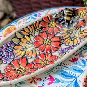 May include: A white ceramic dish with a floral design in shades of red, orange, and yellow. A small bee figurine is attached to the dish.