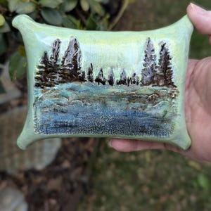 May include: A rectangular ceramic plaque with a light green glazed surface featuring a raised, textured landscape scene. The design depicts dark brown pine trees against a pale sky, with a blue and brown textured foreground representing water.