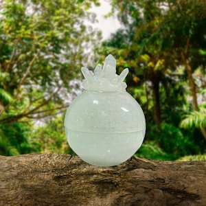 May include: A spherical, light green decorative jar with a crystal-like top, set against a blurred backdrop of lush green foliage. The jar is resting on a piece of weathered wood, creating a natural aesthetic.