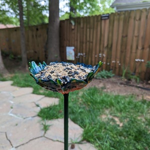 Pu&ograve; includere: Un mangiatoia per uccelli blu e verde a forma di fiore con uno stelo di metallo nero. La mangiatoia &egrave; piena di semi per uccelli.