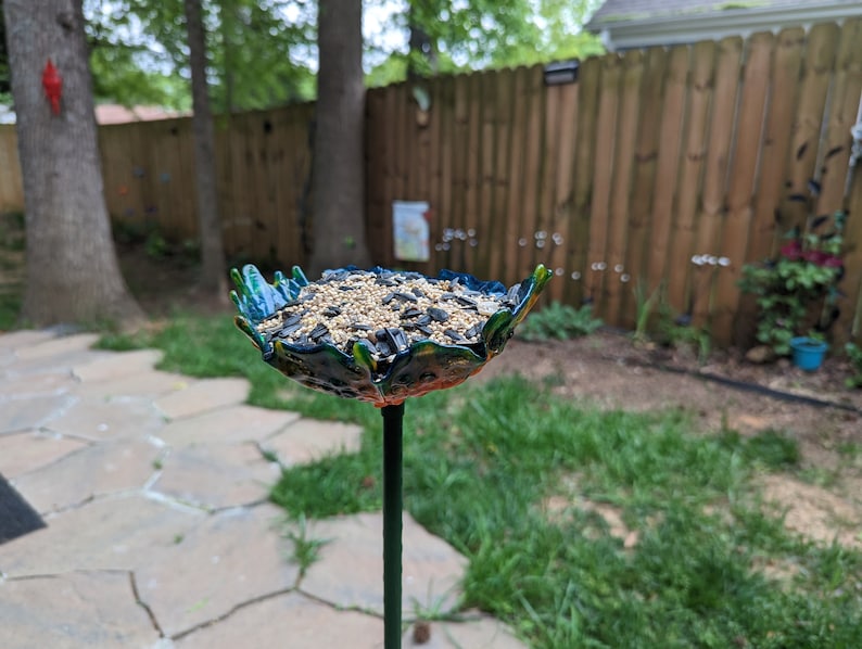 Pu&ograve; includere: Un mangiatoia per uccelli blu e verde a forma di fiore con uno stelo di metallo nero. La mangiatoia &egrave; piena di semi per uccelli.