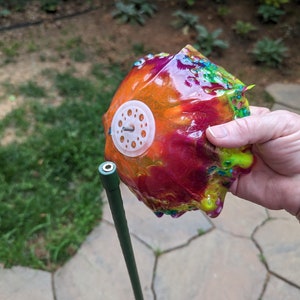 Pu&ograve; includere: Un oggetto rotondo e colorato, rivestito di resina, con un anello di plastica bianca e una vite di metallo al centro. L'oggetto &egrave; tenuto da una mano ed &egrave; attaccato a un palo di metallo verde.