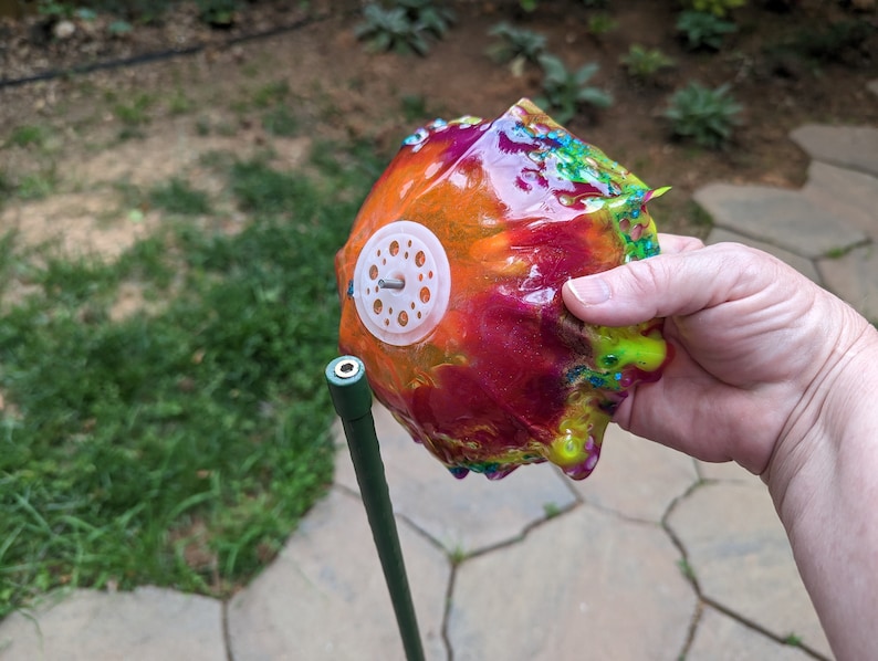 Pu&ograve; includere: Un oggetto rotondo e colorato, rivestito di resina, con un anello di plastica bianca e una vite di metallo al centro. L'oggetto &egrave; tenuto da una mano ed &egrave; attaccato a un palo di metallo verde.