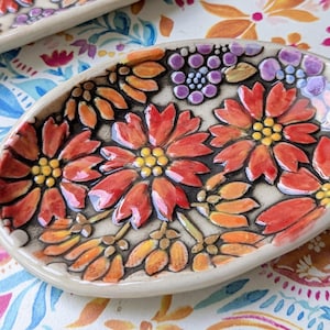 May include: An oval ceramic dish featuring a hand-painted floral pattern with vibrant red, orange, and yellow petals, yellow centers, and small purple clusters. The background is a light cream color with dark outlines defining each flower shape.