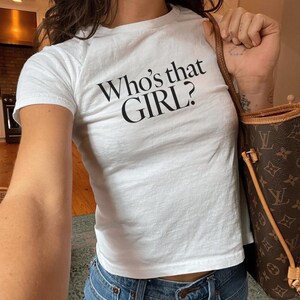 Może przedstawiać: Biała koszulka z nadrukiem "Who's that GIRL?" z przodu. Modelka nosi rozmiar XS.