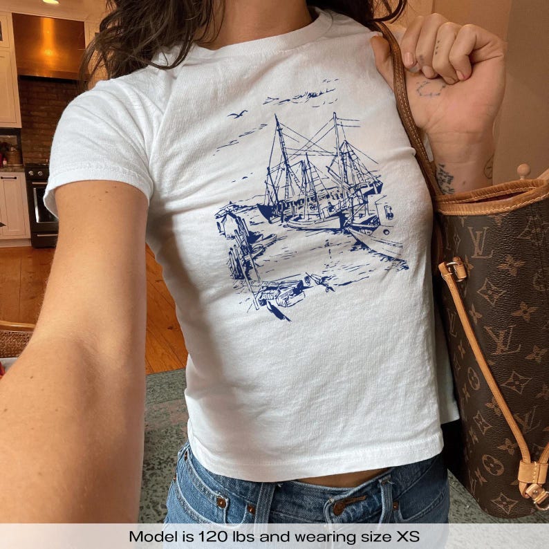 A white t-shirt with a blue graphic of a boat docked at a pier. The graphic is in a vintage style with a simple line drawing. The text 'Model is 120 lbs and wearing size XS, Sailboats Baby Tee, Seashell Baby Tee, Nautical Baby Tee, Nautical T-Shirt, Ocean Theme Crop Tee, Beach Day Crop Top, Summer Baby Tee