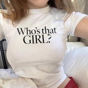 Może przedstawiać: Biały crop top z kr&oacute;tkimi rękawami i nadrukiem "Who's that GIRL?" w kolorze czarnym.