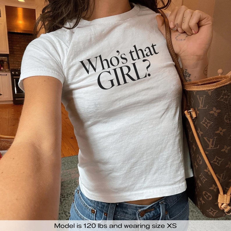Może przedstawiać: Biała koszulka z nadrukiem "Who's that GIRL?" z przodu. Modelka nosi rozmiar XS.
