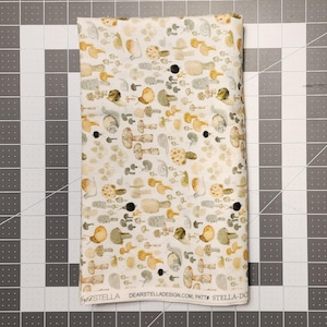 May include: A folded fabric with a repeating pattern of mushrooms in shades of brown, yellow, and gray on a cream background. The fabric has the text "DEARSTELLADESIGN.COM" and "STELLA-DO" printed on it. The fabric is on a gray cutting mat.