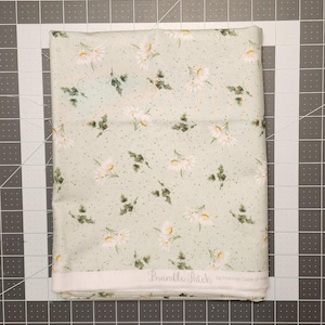May include: A folded fabric with a light green background, featuring a daisy floral pattern with white petals and yellow centers, along with green leaves and small black dots. The text "Bramble Patch" is visible.