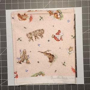 May include: A folded fabric with a light pink background featuring a whimsical pattern of woodland creatures, including rabbits, hedgehogs, and foxes. The fabric has a white border and is decorated with flowers and berries.