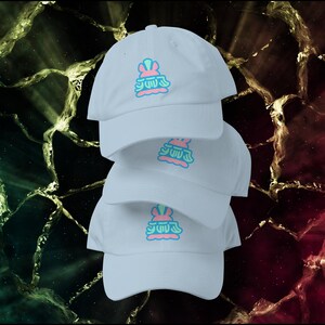 May include: Four light blue baseball caps stacked on top of each other. Each cap has a colorful logo on the front, featuring a pink and blue design. The background has a dark, textured appearance with green, gold, and red tones.