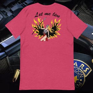 Peut inclure: Un t-shirt rose avec le texte "Let me live" imprimé sur le devant. Le texte est en noir et est entouré d'un motif de flammes jaunes. Une femme avec une lance se tient au milieu des flammes.