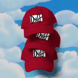 Puede incluir: Una pila de tres gorras de béisbol rojas. Cada gorra presenta la palabra "Duff" en una fuente negra estilizada dentro de un rectángulo blanco. El fondo es un cielo azul con nubes blancas.