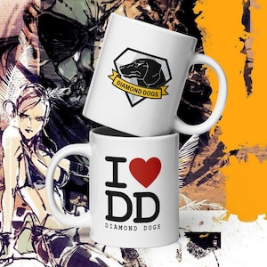 Peut inclure: Deux mugs en céramique blanche. Le mug supérieur présente la silhouette d'un chien noir à l'intérieur d'un losange noir et blanc avec une bannière jaune qui dit "DIAMOND DOGS". Le mug inférieur a "I ♥ DD" avec un cœur rouge et les mots "DIAMOND DOGS".