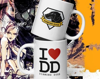 May include: Two white ceramic mugs. The top mug features a black dog silhouette inside a black and white diamond with a yellow banner that reads "DIAMOND DOGS". The bottom mug has "I ♥ DD" with a red heart and the words "DIAMOND DOGS".