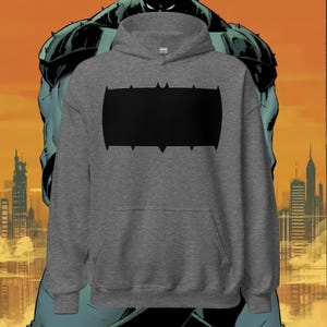 Puede incluir: Una sudadera con capucha gris con un diseño gráfico rectangular negro con bordes puntiagudos. La sudadera tiene un bolsillo delantero y una capucha con cordón. El fondo presenta una ilustración de cómic de un superhéroe y un horizonte urbano.