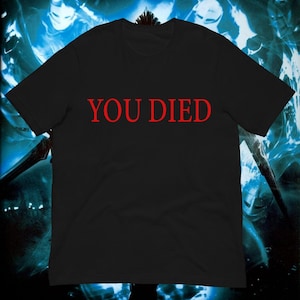 May include: A black short-sleeved t-shirt featuring the text YOU DIED printed in a bold red serif font across the center of the chest. The garment has a classic crew neckline and a straight hemline, presenting a simple and clean design.