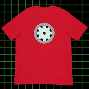 Puede incluir: Una camiseta roja de manga corta con un gráfico central de un emblema circular. El emblema consiste en una forma de engranaje de color verde menta claro con ocho muescas negras, rodeado por un borde circular de color violeta claro sobre fondo rojo.