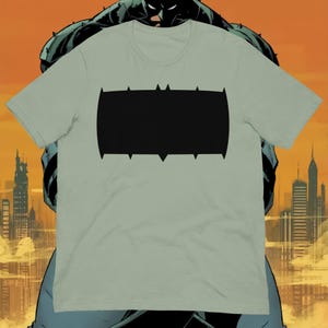 May include: A light green t-shirt featuring a large black emblem. The emblem is a horizontal rectangle with pointed edges. The shirt is a short-sleeved crew neck style. The background features a comic book illustration of a city skyline and a superhero.