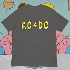 Peut inclure: Un t-shirt gris anthracite à manches courtes avec le nom du groupe AC/DC imprimé en lettres jaunes audacieuses sur la poitrine. Le motif comprend un symbole d'éclair jaune positionné entre les lettres AC et DC sur le devant du vêtement.