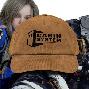 May include: A brown corduroy baseball cap with the text "CABIN SYSTEM" in black. The cap features a smiling face graphic on the left side of the text. The hat is a classic six-panel design with a curved brim.