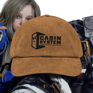 May include: A brown corduroy baseball cap with the text "CABIN SYSTEM" in black. The cap features a black graphic of a smiling box. The hat is a classic six-panel design with a curved brim.