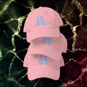 May include: Four pink baseball caps stacked on top of each other. Each cap has a curved brim and a pastel blue and pink embroidered design. The background is a mix of green, black, and red.