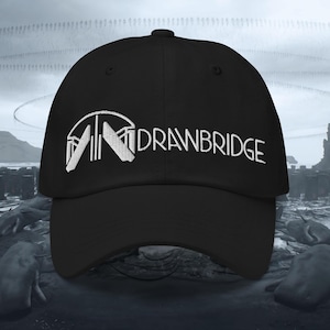 May include: A black baseball cap featuring white embroidery on the front panel. The design includes a stylised graphic of a bridge structure on the left and the text "DRAWBRIDGE" in a clean, sans-serif font extending to the right side.