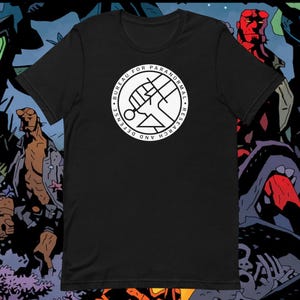 Op de afbeelding: Zwart T-shirt met een witte cirkelvormige afbeelding met de tekst "BUREAU FOR PARANORMAL RESEARCH AND DEFENSE".