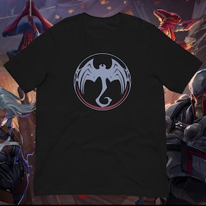 May include: Black t-shirt featuring a circular design with a stylized dragon or bat-like creature in light blue, outlined in red and gray. The graphic is centered on the front of the shirt. The shirt is made of a soft material.