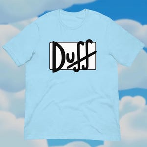 Op de afbeelding: Lichtblauw T-shirt met het woord "Duff" in zwarte, gestileerde tekst in een zwarte rechthoek. Het shirt is gemaakt van een zacht materiaal en heeft korte mouwen en een ronde hals. De achtergrond is een blauwe lucht met witte wolken.