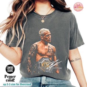 Può includere: Una t-shirt grigio antracite a maniche corte con una stampa grafica di Charles Oliveira con una cintura da campionato UFC. Il design include una firma bianca e il testo Charles Oliveira. La maglia presenta un girocollo.