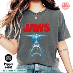Puede incluir: Una camiseta de manga corta gris carbón con el texto rojo JAWS sobre un gráfico de un nadador en la superficie del agua y un gran tiburón con la boca abierta emergiendo de las profundidades del agua azul. Tiene cuello redondo.