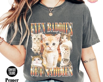 Puede incluir: Una camiseta gris carbón con un estampado gráfico de cinco gatos con expresiones tristes. El texto "EVEN BADDIES GET SADDIES" está impreso en una fuente negrita con degradado de naranja a amarillo. El diseño incluye rayos detrás de los gatos.