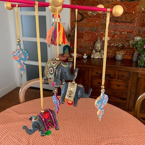 Peut inclure: Un mobile en bois avec quatre ornements suspendus. Les ornements sont un motif cachemire bleu et vert, un éléphant gris avec des embellissements roses et dorés, un éléphant gris avec des embellissements dorés et blancs, et un éléphant gris avec des embellissements roses, dorés et rouges.