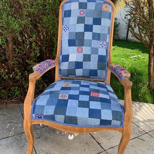 Peut inclure: Fauteuil vintage avec une structure en bois brun clair. L'assise et le dossier sont recouverts d'un patchwork de carrés de denim bleu, rehaussés de patchs brodés colorés. Les accoudoirs présentent un tissu à motifs.