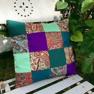 Peut inclure: Un coussin patchwork avec un motif paisley vert, violet et marron. Le coussin est posé sur une chaise en métal blanc.
