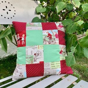 Peut inclure: Un coussin patchwork rouge et vert avec une variété d'illustrations vintage, notamment des enfants à vélo, une fille regardant par une fenêtre et un livre avec le texte "Le livre est un ami" dessus.