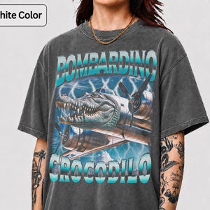 Op de afbeelding: Een grafietkleurig t-shirt met korte mouwen en een grafische print van een alligatorhoofd geïntegreerd in een vintage vliegtuig. De tekst "BOMBARDINO" staat boven de afbeelding en "CROCODILO" eronder, beide in een blauw verloop.