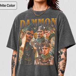 以下が含まれることがあります： グラファイトカラーのTシャツ。角と尖った耳を持つファンタジーキャラクターのヴィンテージスタイルのグラフィックが特徴です。「DAMMON」の文字が金色でプリントされています。シャツは使い古されたような外観をしています。